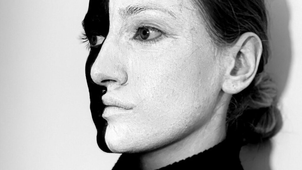 A woman in three quarters profile wearing a black turtleneck and face paint that covers her face in half white and half black paint.