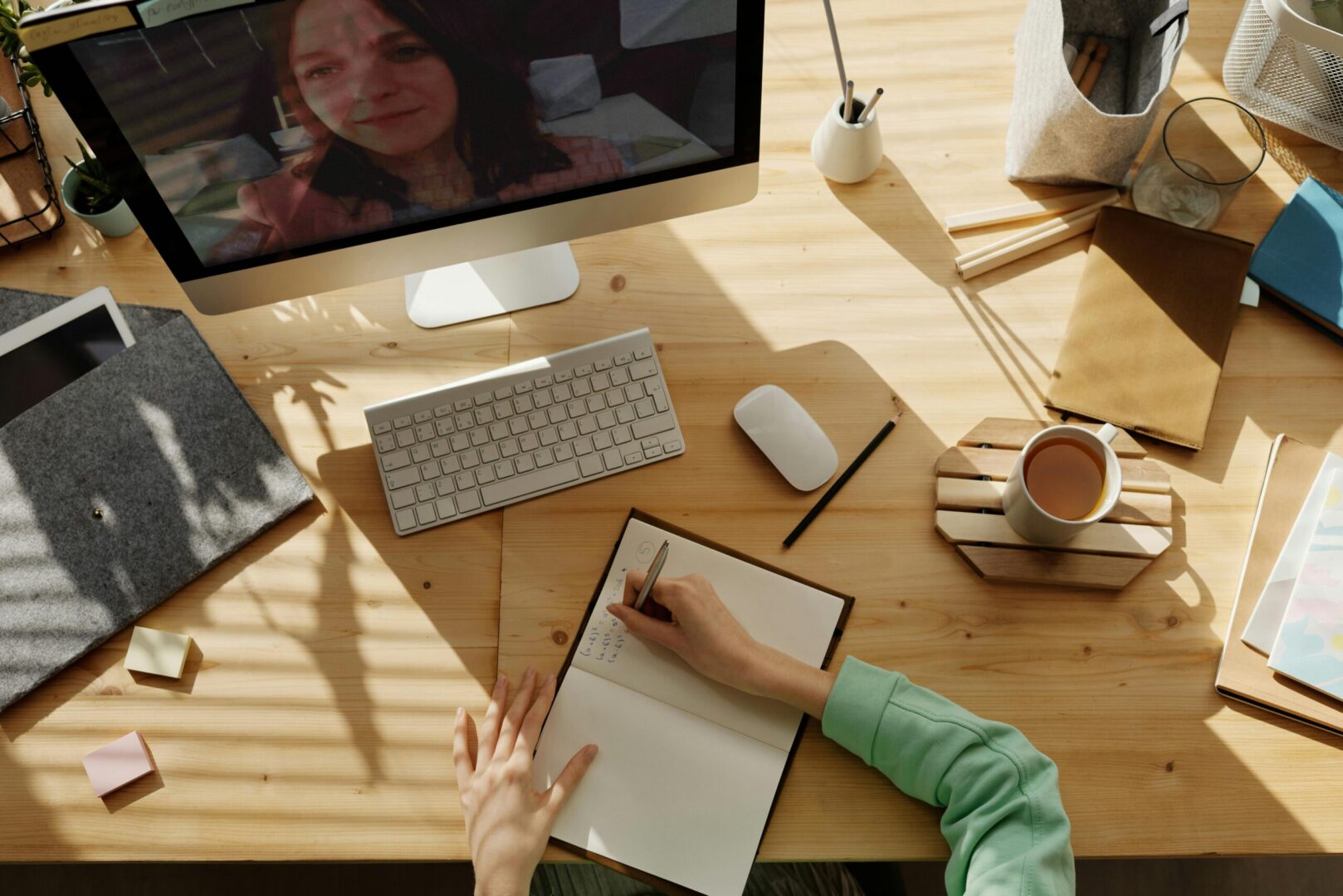 Person doing math in a notebook on a desk with various stationary items and a computer screen showing live virtual instruction