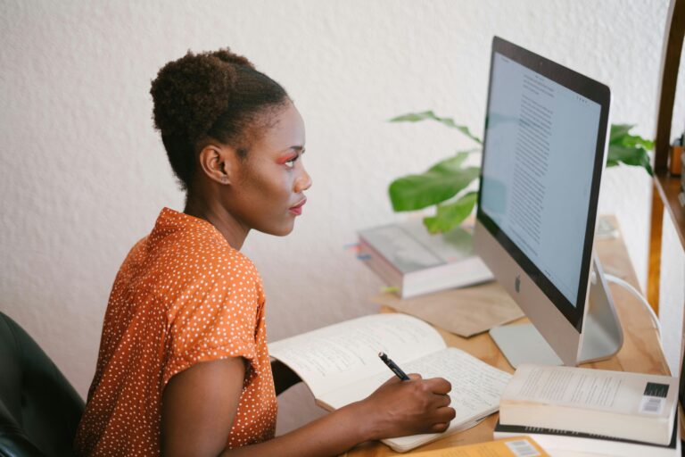 woman looking at desktop computer screen and writing on paper.