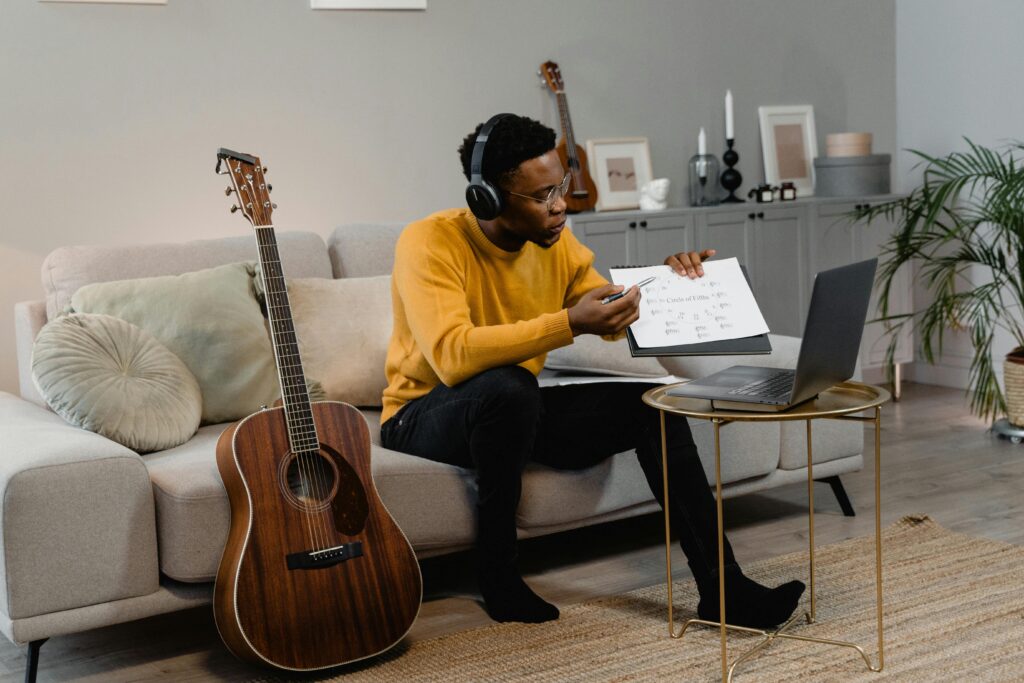 man sitting on couch with a guitar and holding a paper in front of a laptop